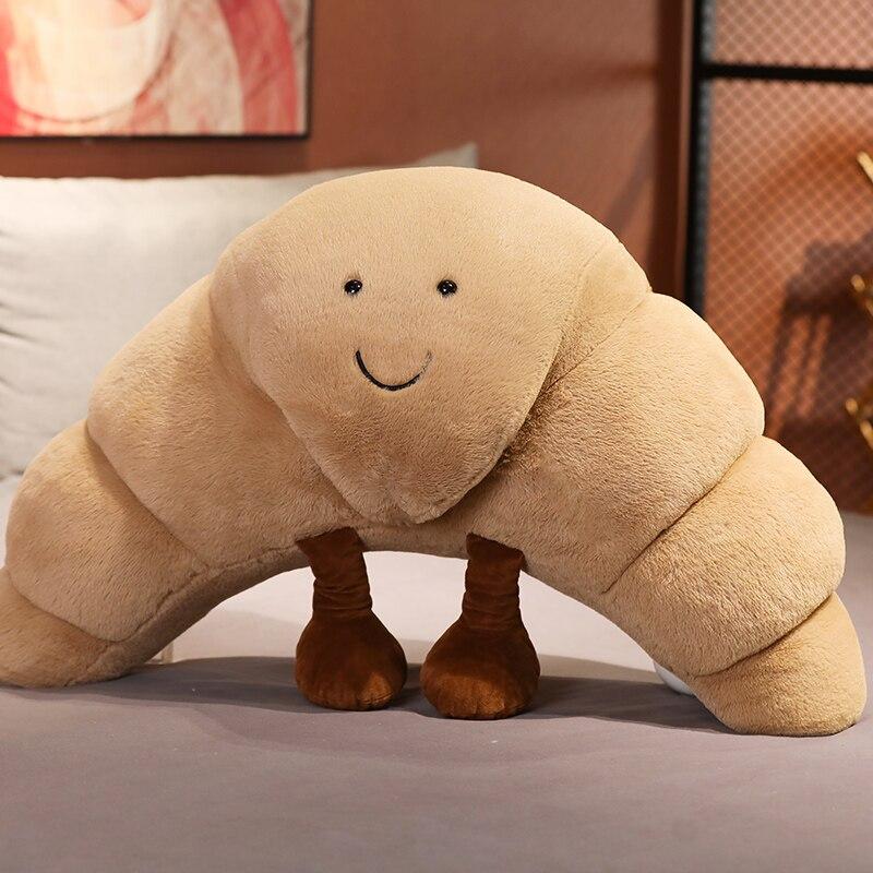 Cute beige croissant plush toy with smiling face, soft PP cotton filling, and small brown feet. Available in three sizes.